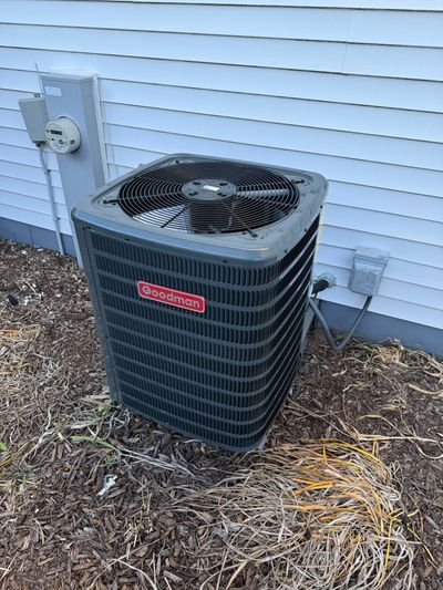 A gray residential air conditioning unit sitting outside against a white siding wall surrounded by brown mulch.