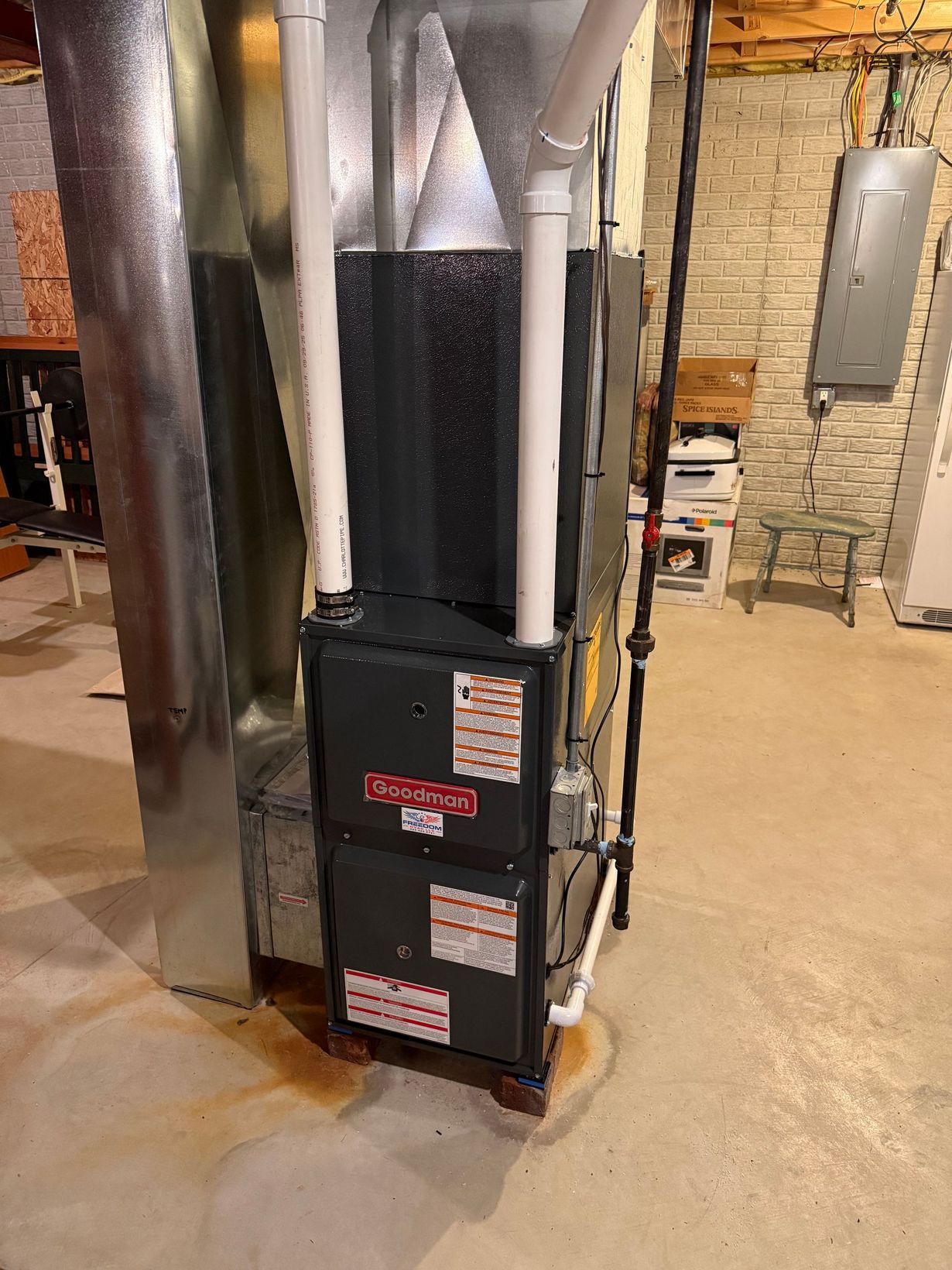 A black Goodman high-efficiency furnace stands on a basement floor next to a large metal duct and white PVC pipes.