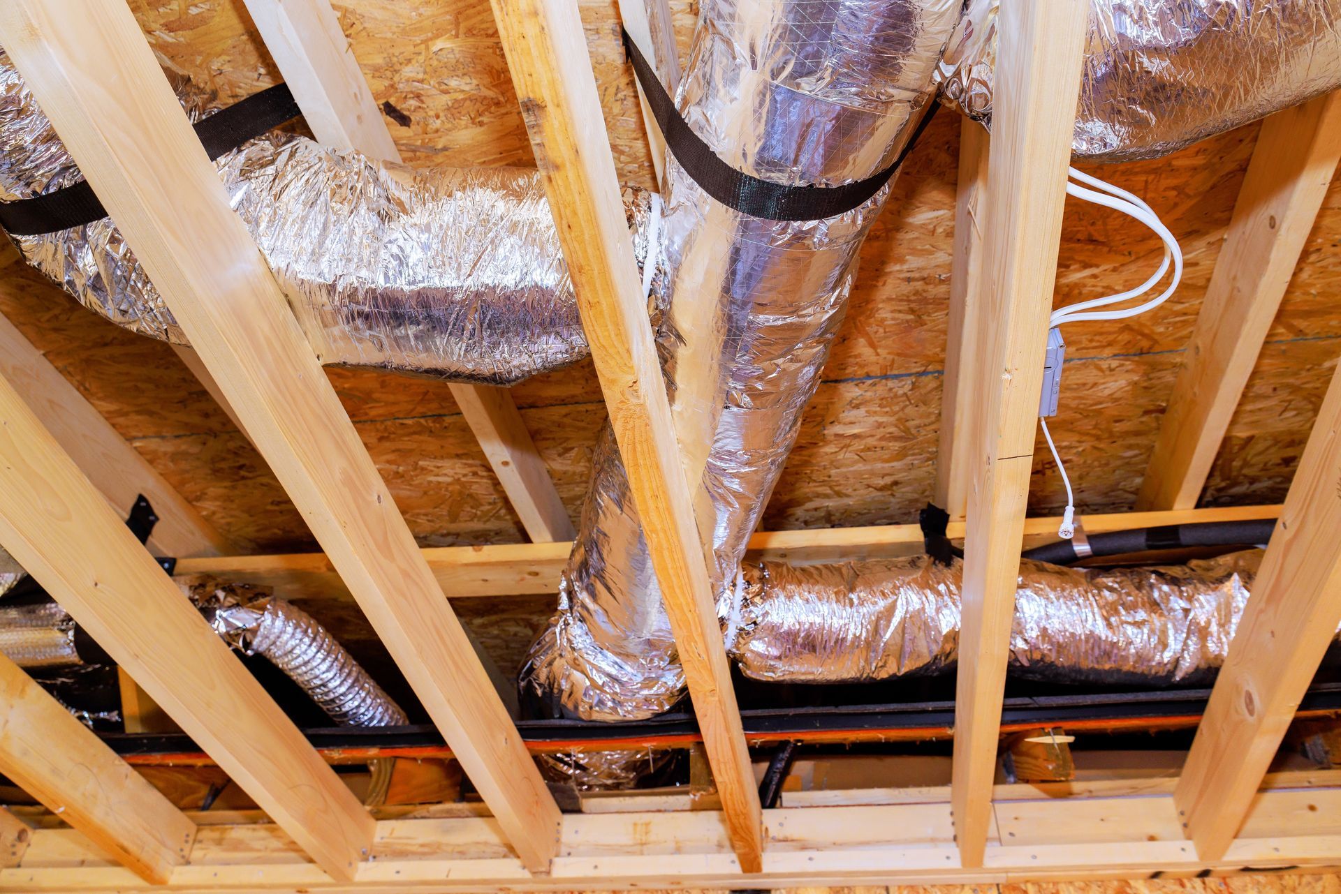 Insulated foil flexible HVAC ducts running between wooden ceiling joists in an unfinished attic space.