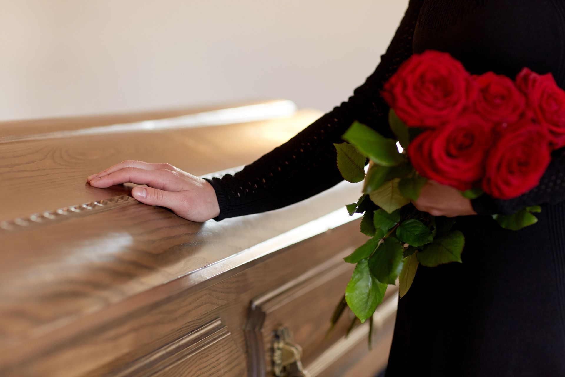 Person in black attire holding red roses beside a polished wooden casket