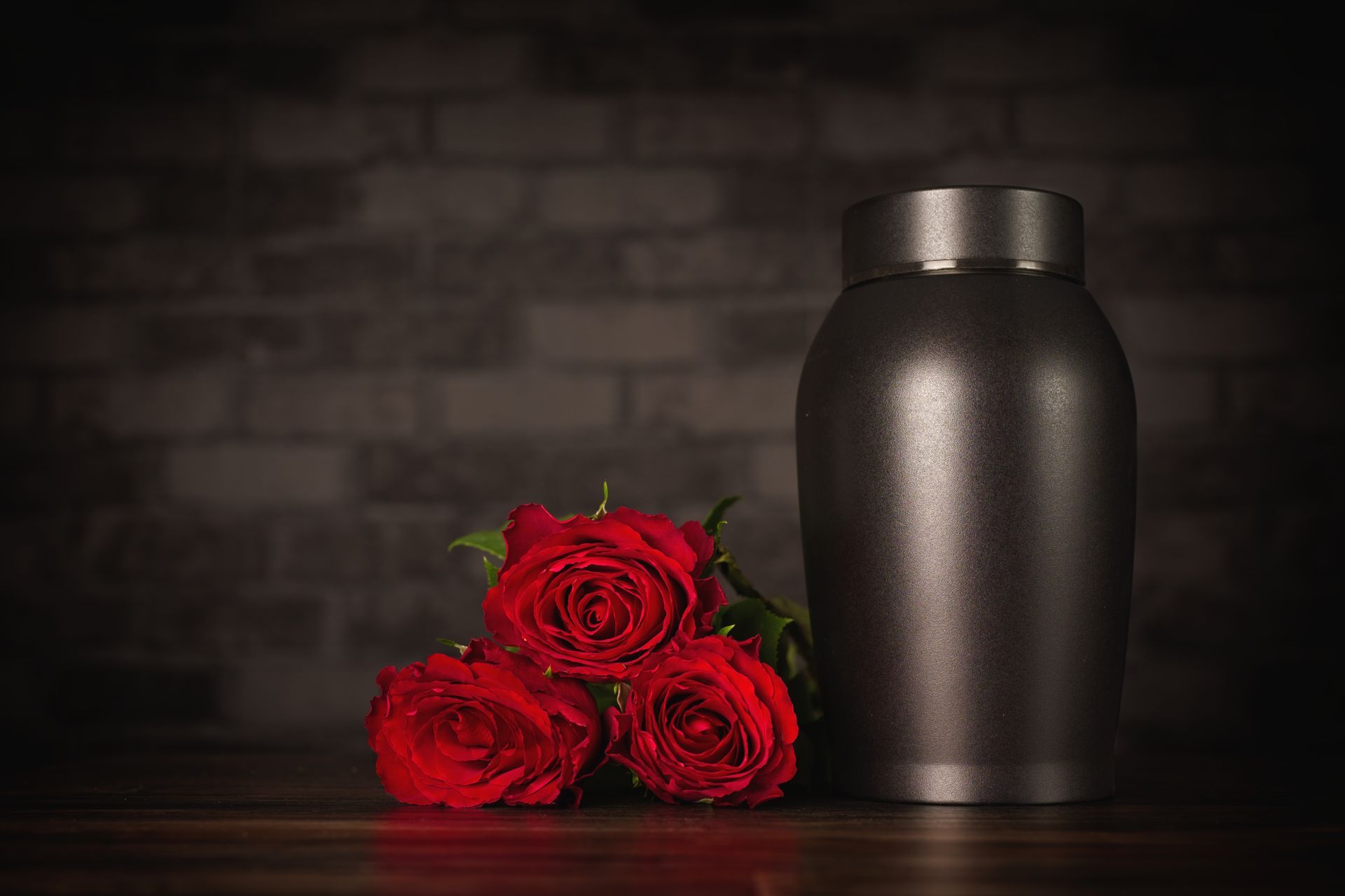 Red roses beside a matte black urn on a dark tabletop, against a blurred brick wall.