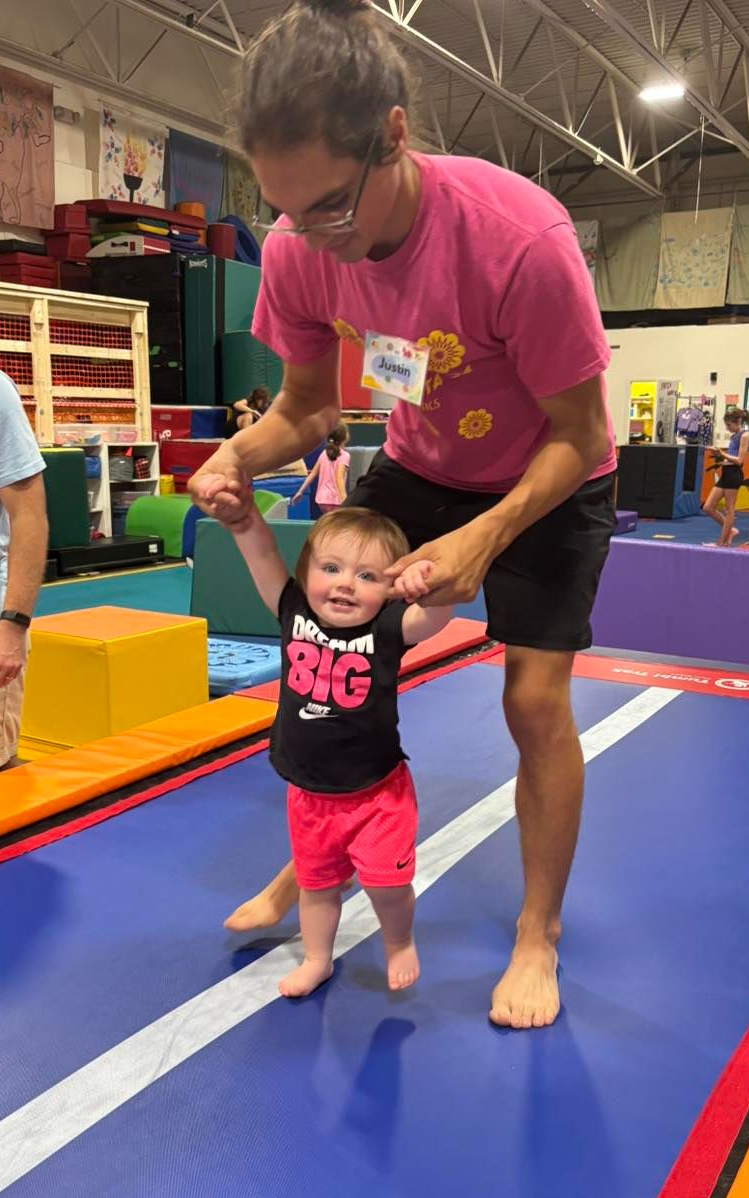 A man helps a smiling baby take steps on a blue mat in a gym. The baby wears a black 