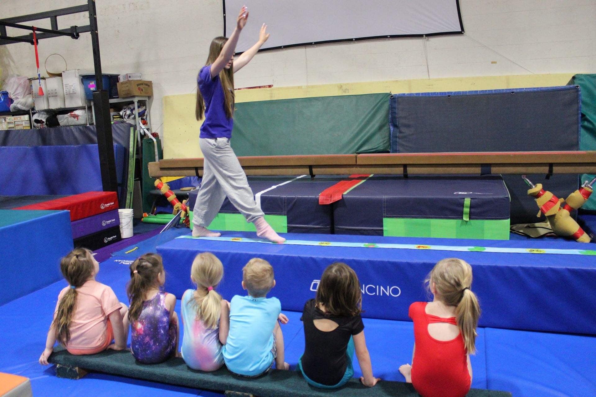 A young instructor balances on a beam, while six young girls watch from a mat in a gym.