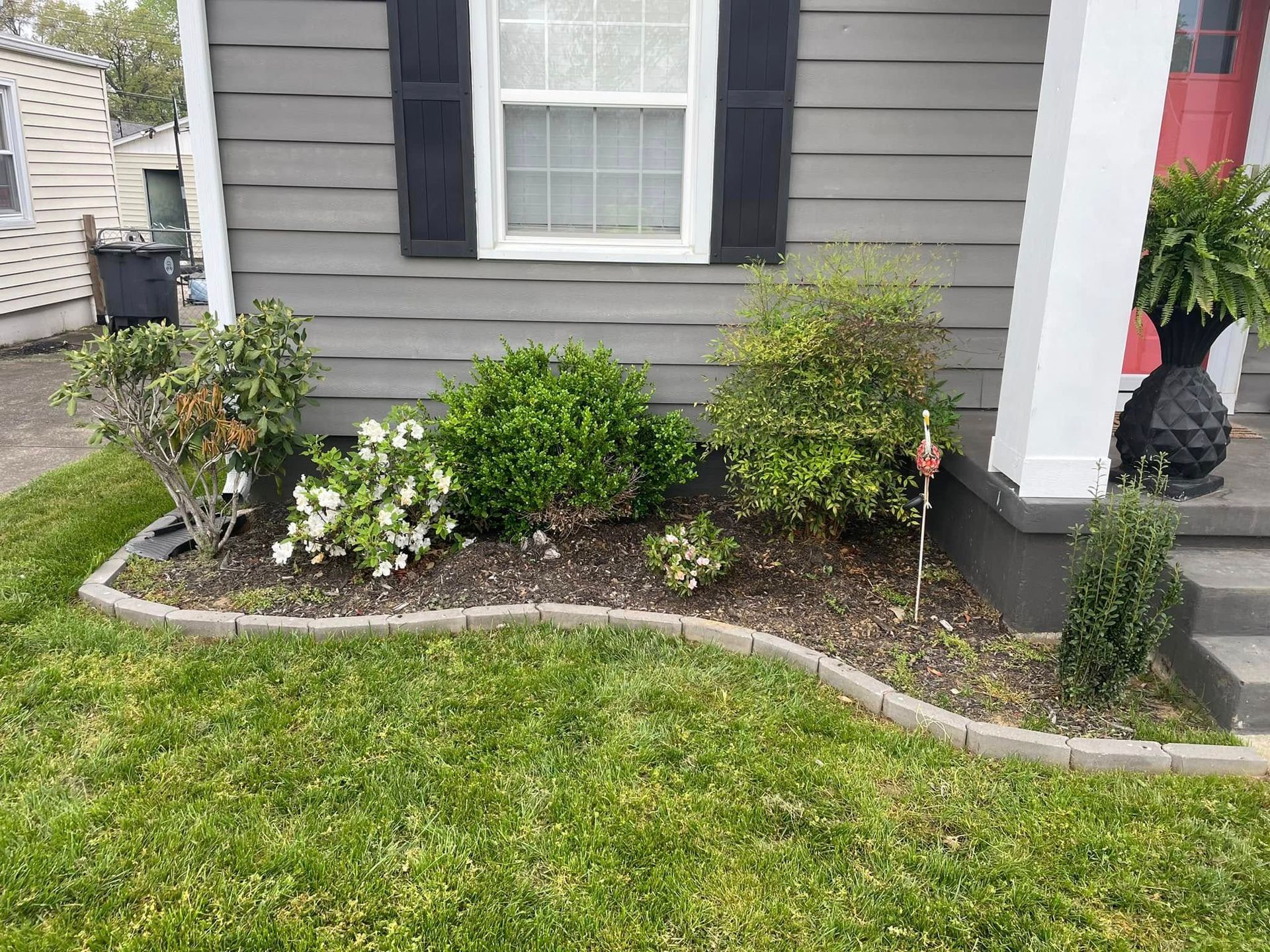 A landscaped front yard bed with green shrubs, flowers, and a brick border near a gray house.