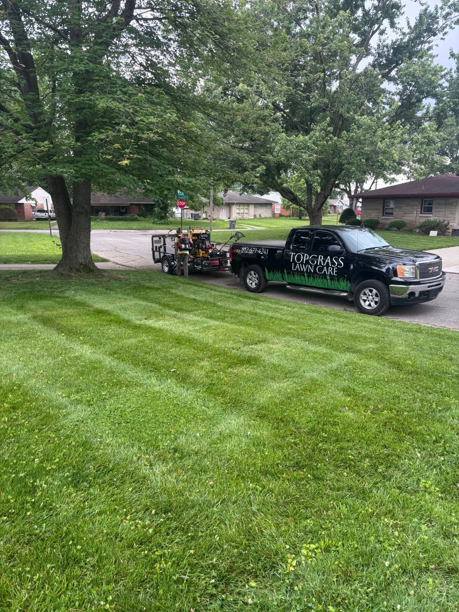 Black truck with lawn care equipment parked on green grass near a tree and houses.