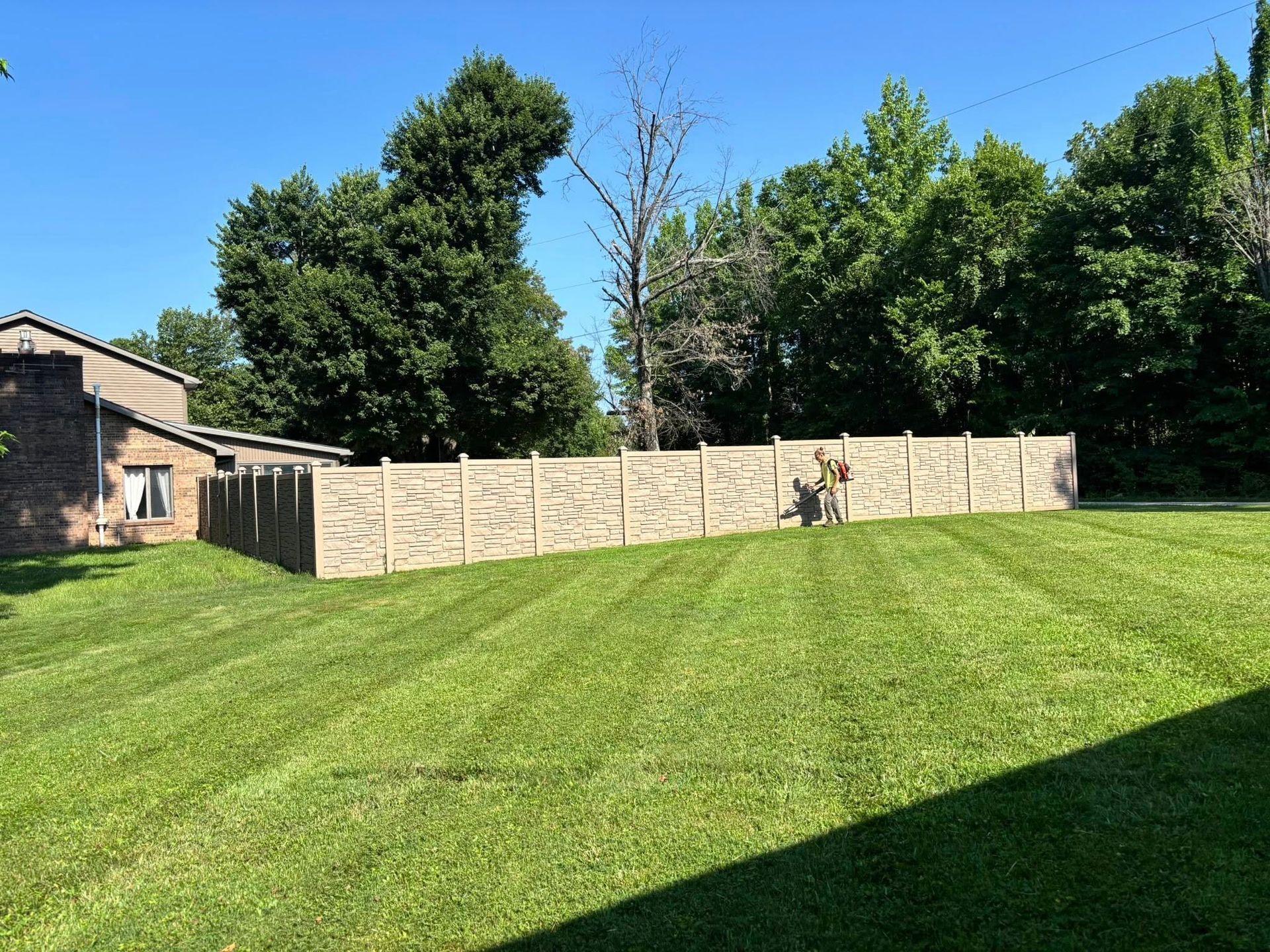 A brick-patterned fence on a grassy hill in front of trees and a house on a sunny day.