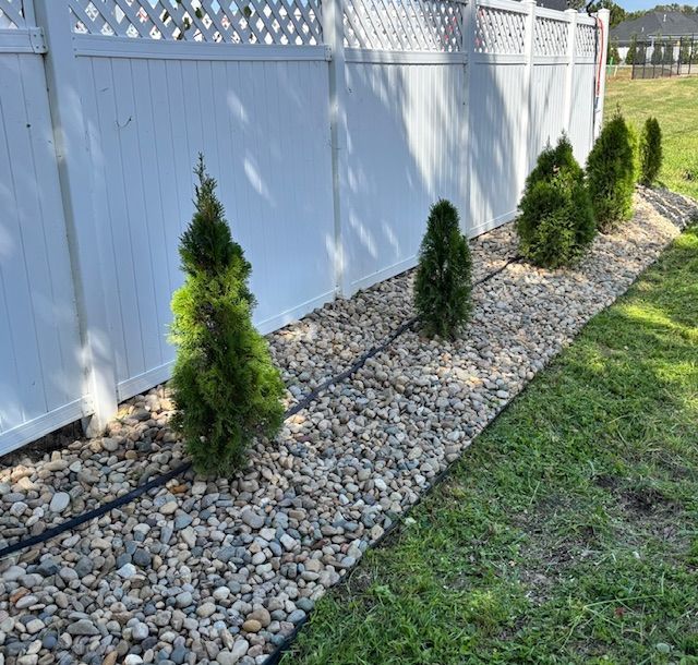 Person mulching garden with wheelbarrow and trailer; green plants, brown mulch, and grey pavement.
