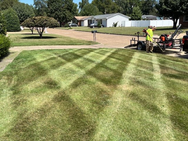 A person on a yellow riding lawnmower cuts a striped pattern into a green lawn in a backyard.
