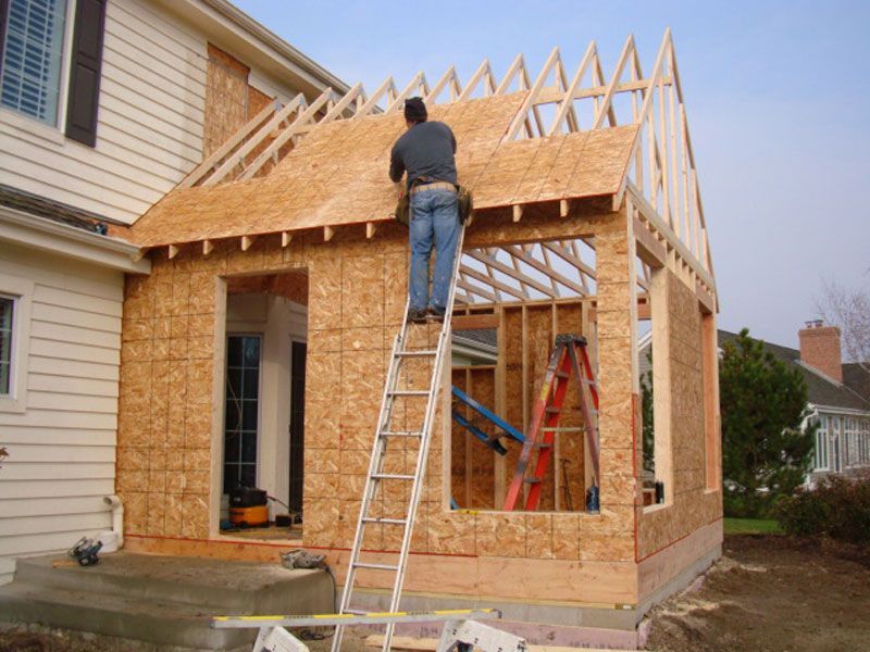 Man on ladder building an addition to a house, attaching plywood to the roof frame.