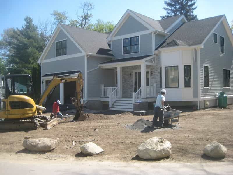 Construction site in front of a gray house. Workers use machinery; dirt and rocks are on the ground.