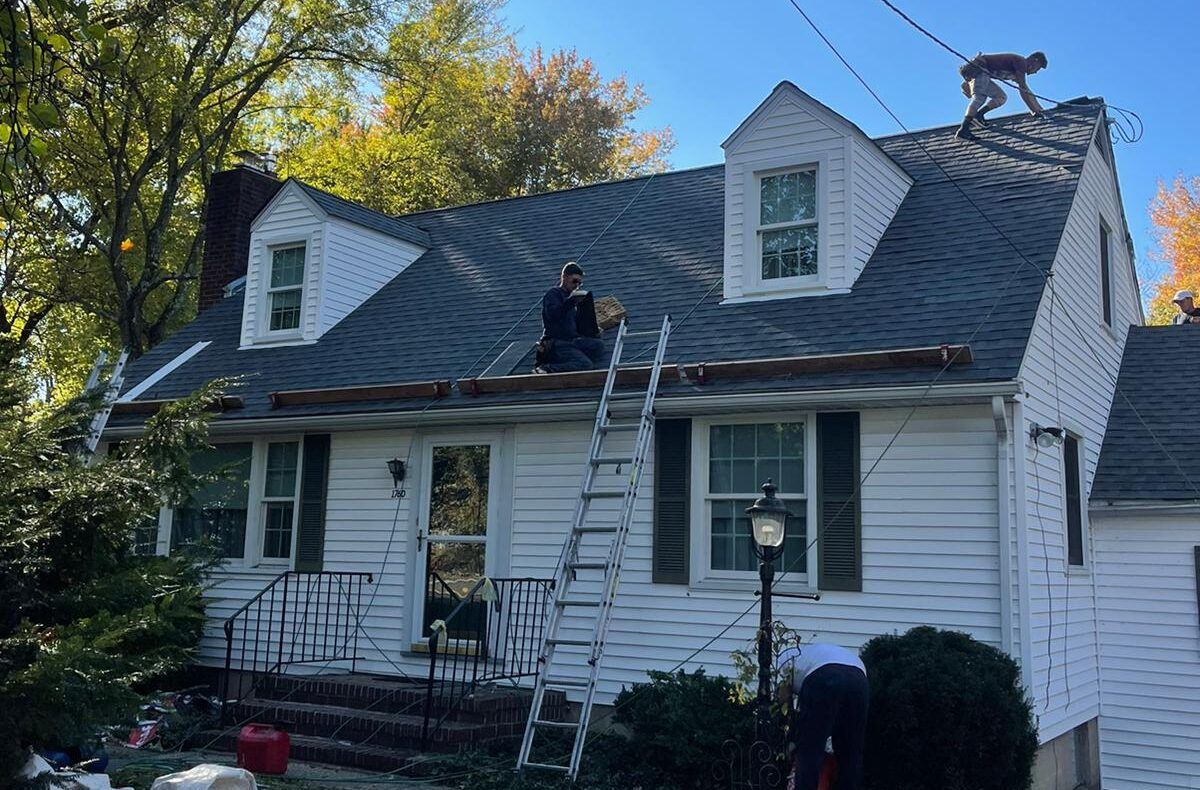 Workers on a roof of a white house with a ladder. One on the rooftop, others working at the edge.