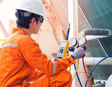 Technician in orange coveralls checks HVAC system with gauges, outdoors.