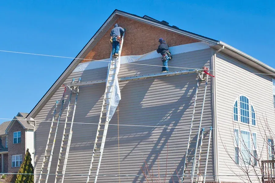 Two roofers working on the side of a house, using ladders and scaffolding on a sunny day.