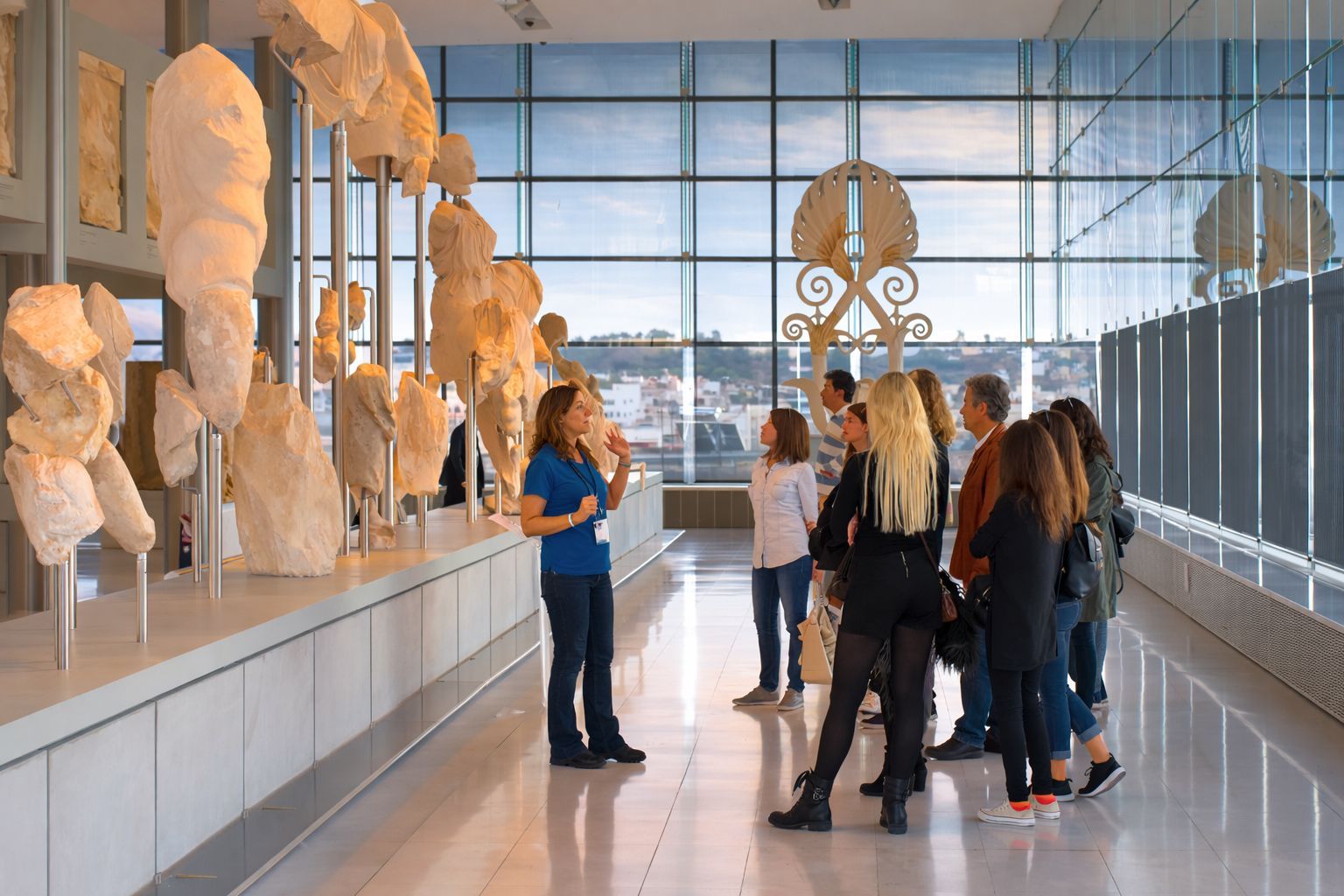 A guide speaks to a group of visitors in a bright, modern museum gallery featuring ancient stone sculptures on display.