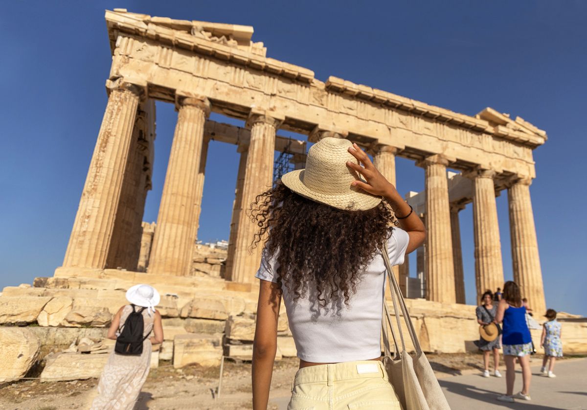 A person wearing a straw hat and white top stands before the Parthenon in Athens with other visitors nearby.