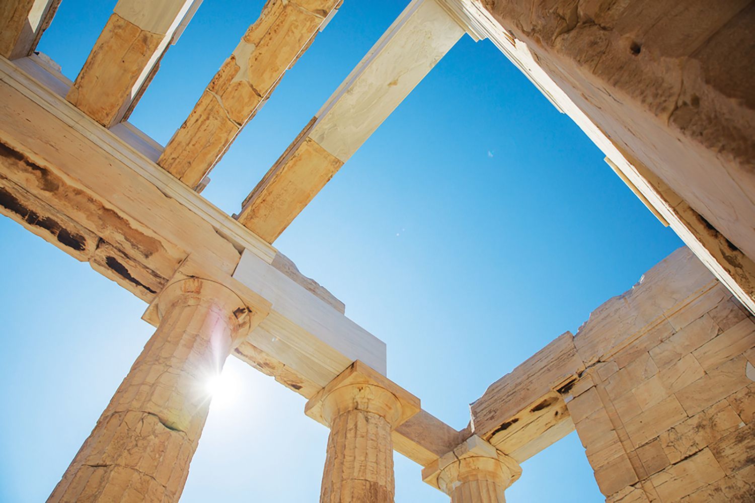Low-angle view of ancient Greek temple ruins with stone columns framing a bright blue sky.
