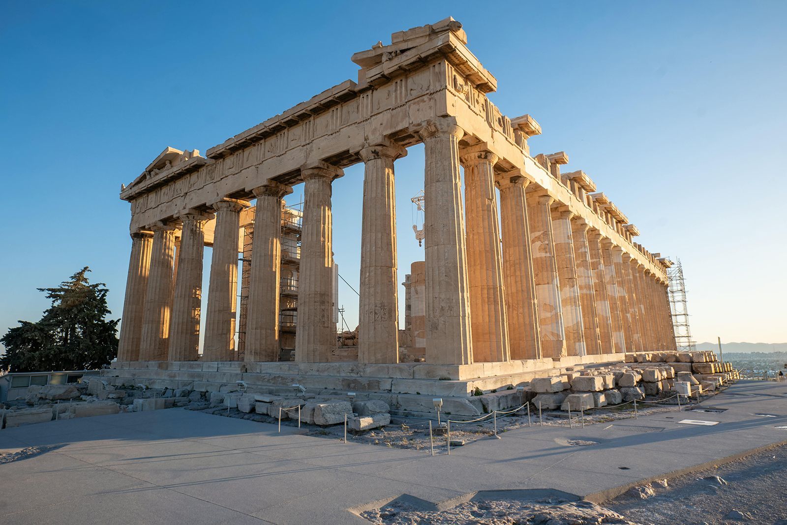 The Parthenon, an ancient Greek temple with weathered columns, stands on a rocky hill under a clear blue sky.