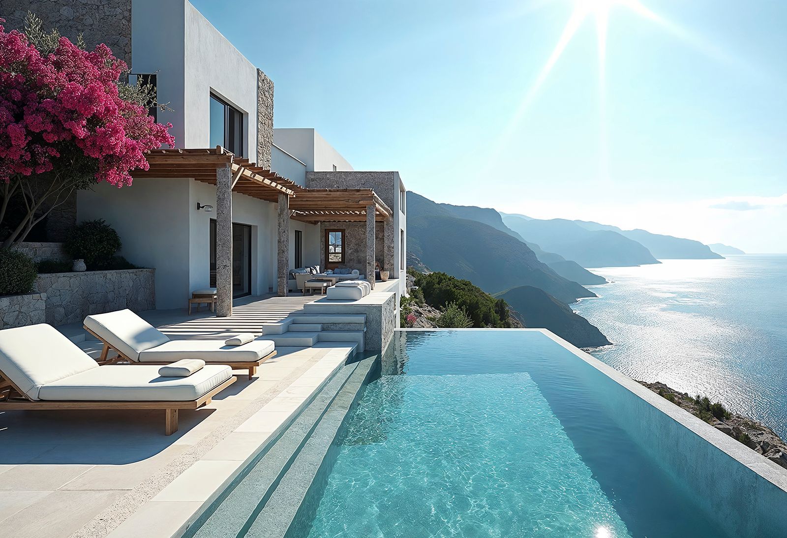 An infinity pool overlooks a sunlit Mediterranean coastline beside a white villa with stone accents and bougainvillea.