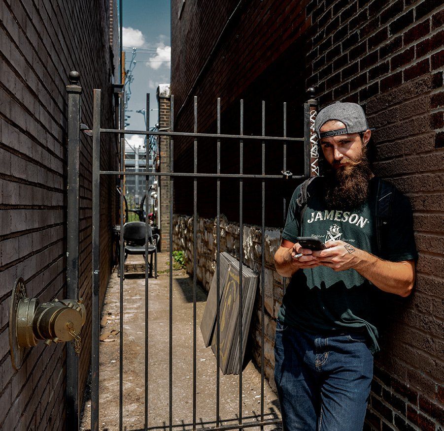 a bartender on break wearing a Jameson whiskey shirt leans against a brick wall during his break at a local bar in The Loop, a revitalized shopping and entertainment area in University City, Missouri.