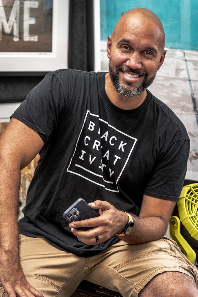 An artist exhibiting in the 2023 Clayton Art Fair in St. Louis, Missouri, wearing a black t-shirt is sitting on a bench holding a cell phone . 