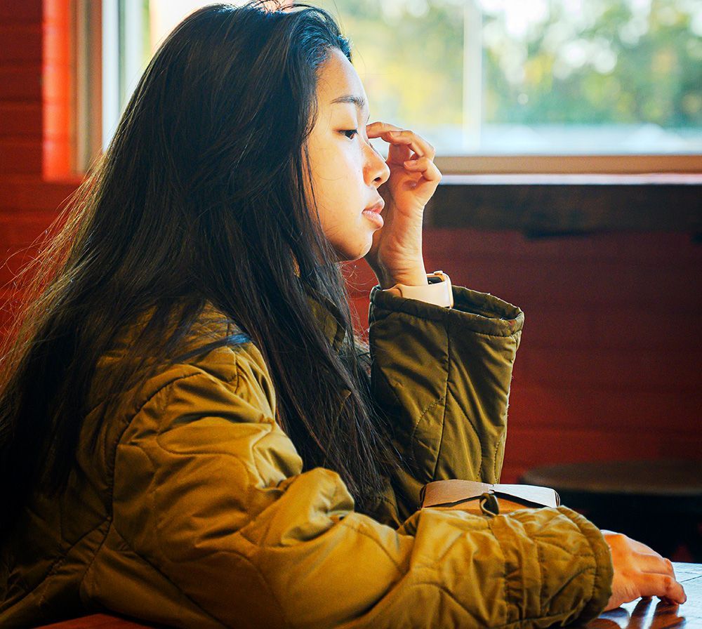 a woman in a tan jacket wipes her eyes with her hand in a coffee shop while waiting for her order. 