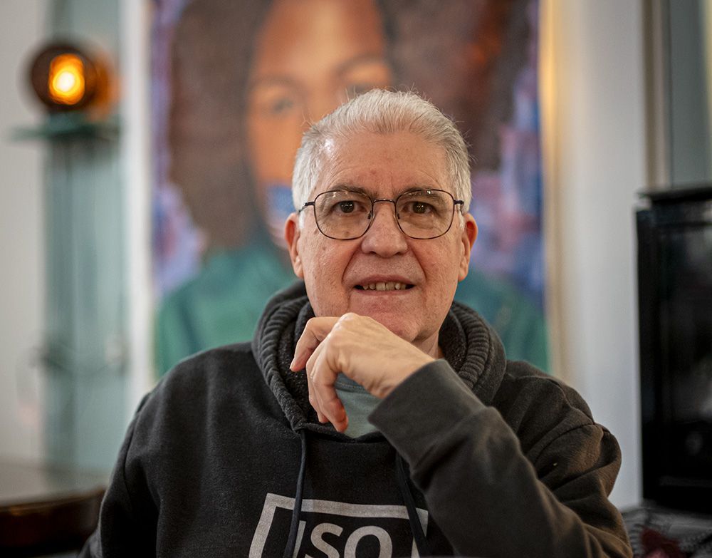 Chuck Ramsay,photographer,  wearing glasses and a hoodie and sitting in front of a painting. Photo by John Montre using Chuck's Leica M10 and Voightlander 35mm f1.2 lens in The Drip Community Coffee House, St. Louis, MO.  Chuck is a retired corporate communications professional who served as the Communications Director for the Bar Association of Metropolitan St. Louis prior to his retirement in 2015, among other companies and industries for over 45 years.