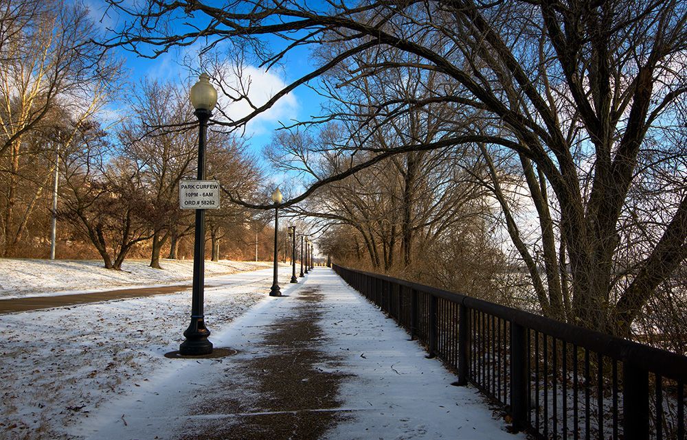 a snowy path in a park with a sign that states park curfew hours, Sister Marie Charles Park in Carondelet neighborhood of St. Louis, along the banks for the Mississippi River, in sub-zero weather.
