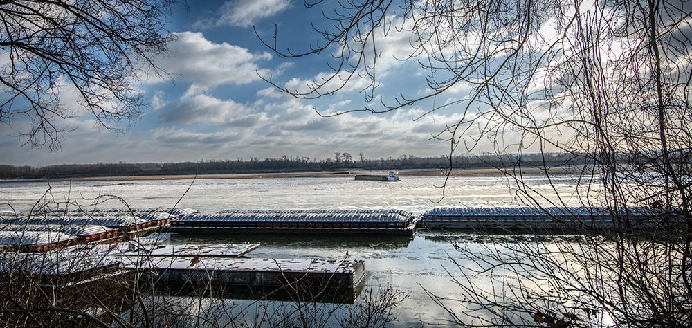 a large body of water with trees in the foreground and a boat in the background. Tugboat and barges on the Mississippi River in winter. Ice on the river, sub-zero morning from the western bank of the river in Sister Marie Charles Park in the Carondelet neighborhood of St. Louis, Missouri.