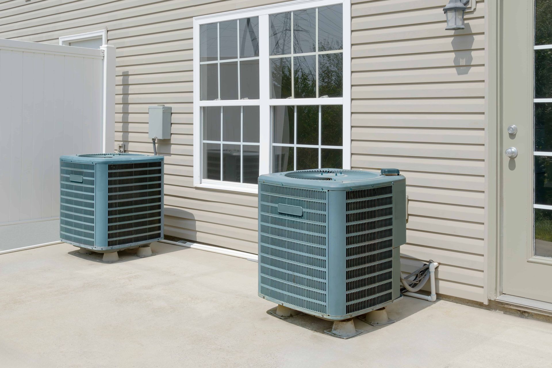 Two air conditioners are sitting outside of a house next to a window.
