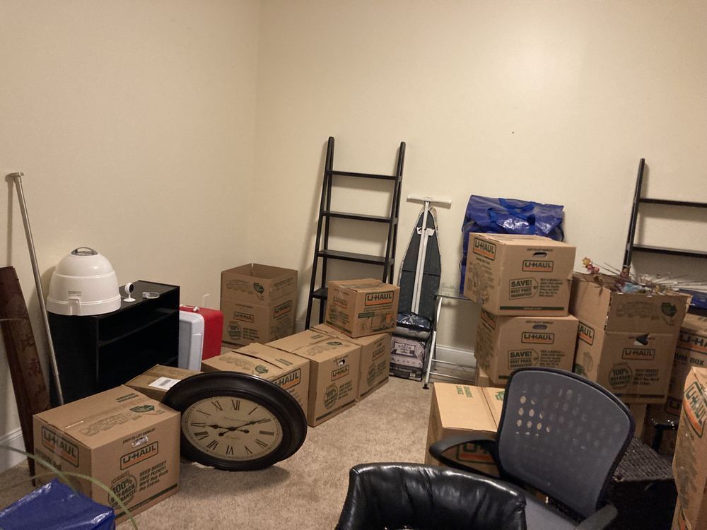Room filled with cardboard boxes, a clock, ladder, and ironing board, indicating a move or storage.