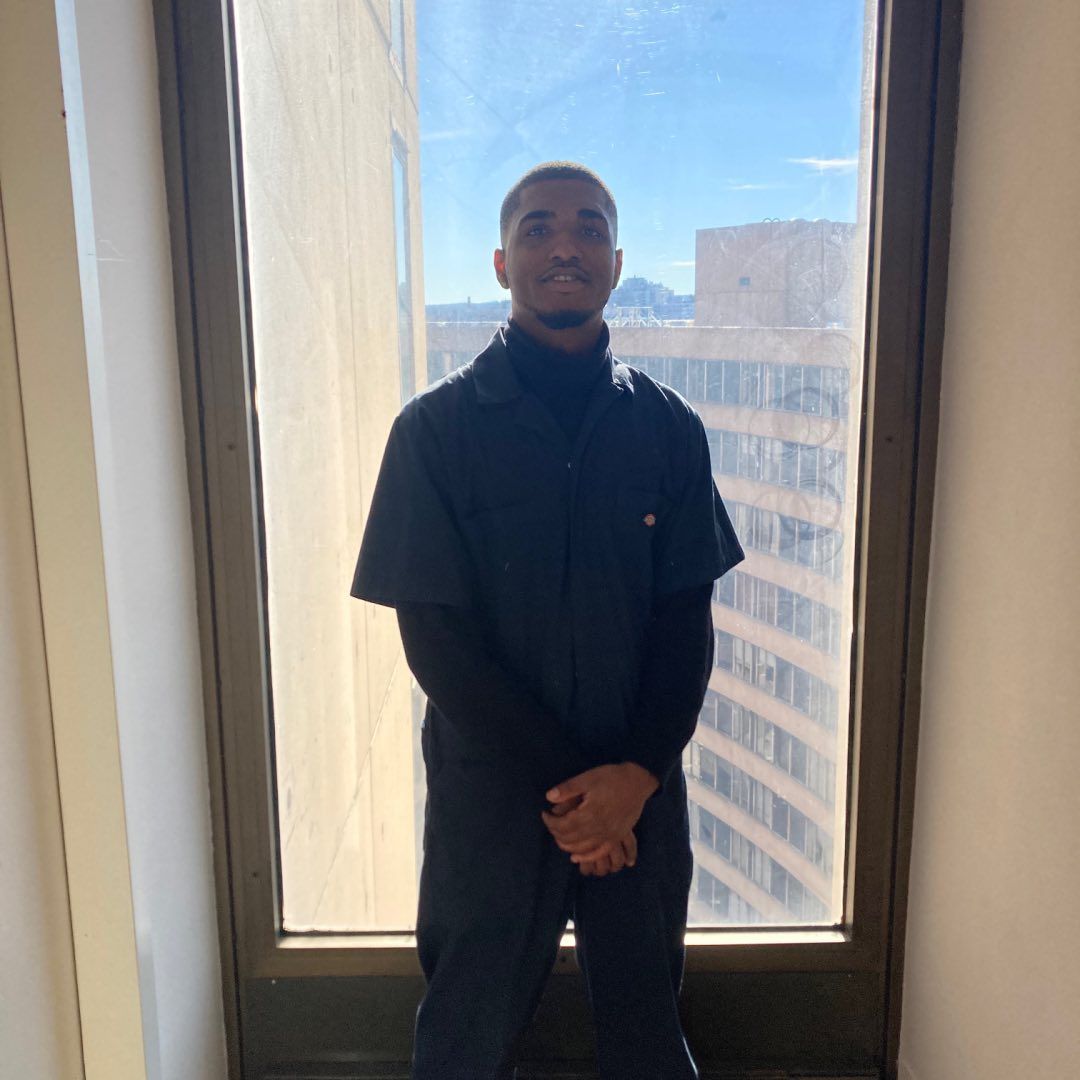 Man in black turtleneck and jumpsuit standing in front of a window, city view.