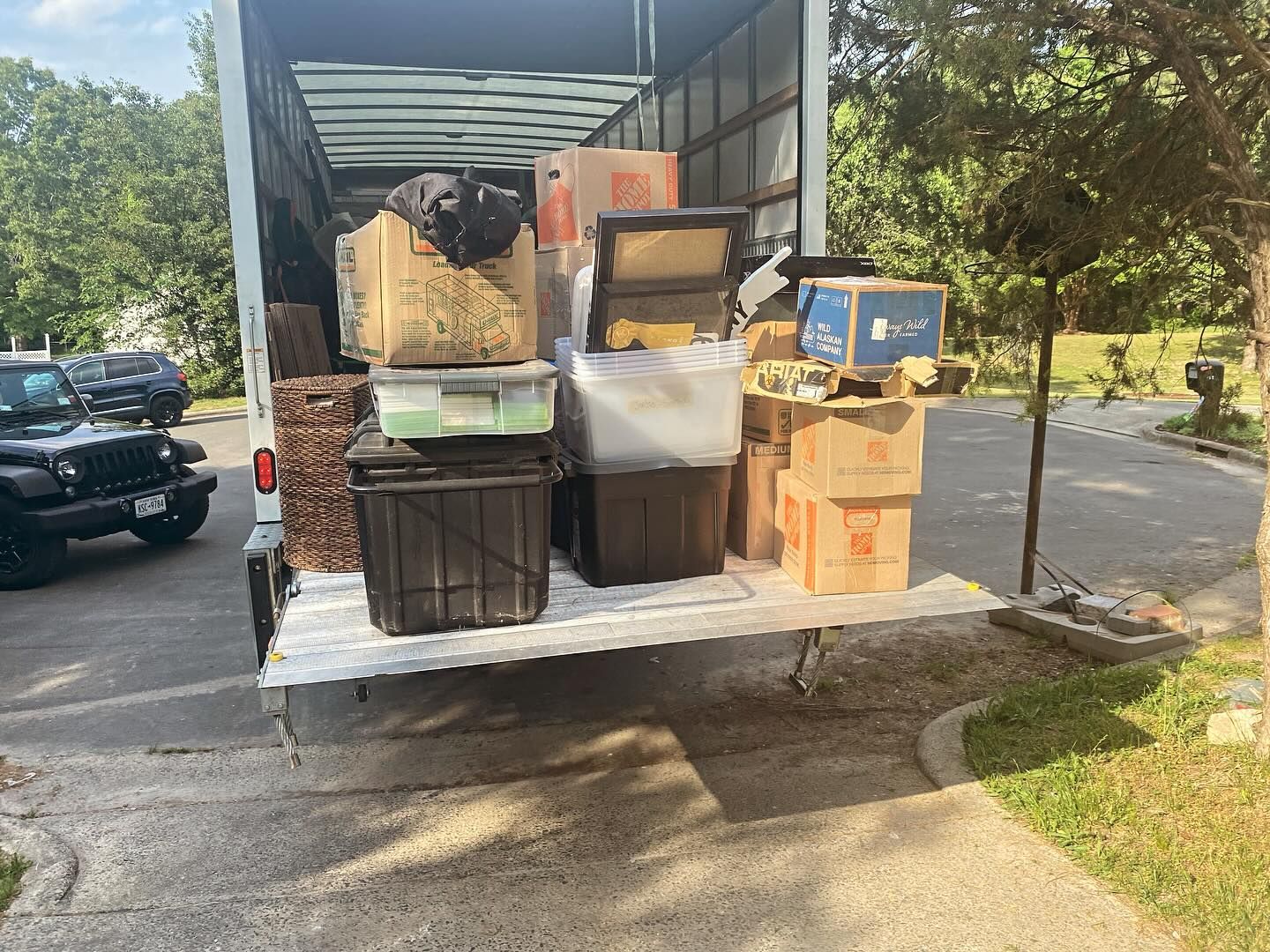 Truck bed filled with moving boxes and bins; parked outside, near a green jeep.