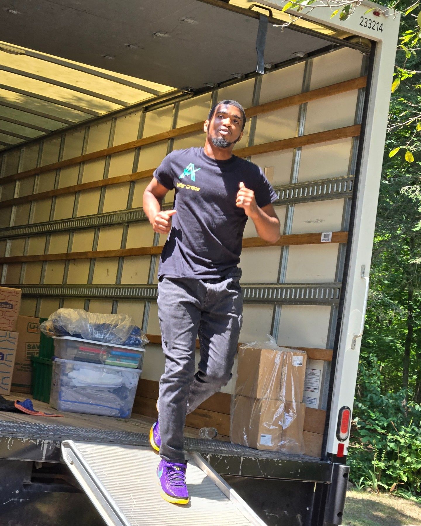 Man running out of a moving truck onto a ramp, boxes inside. He wears a dark t-shirt and purple shoes.