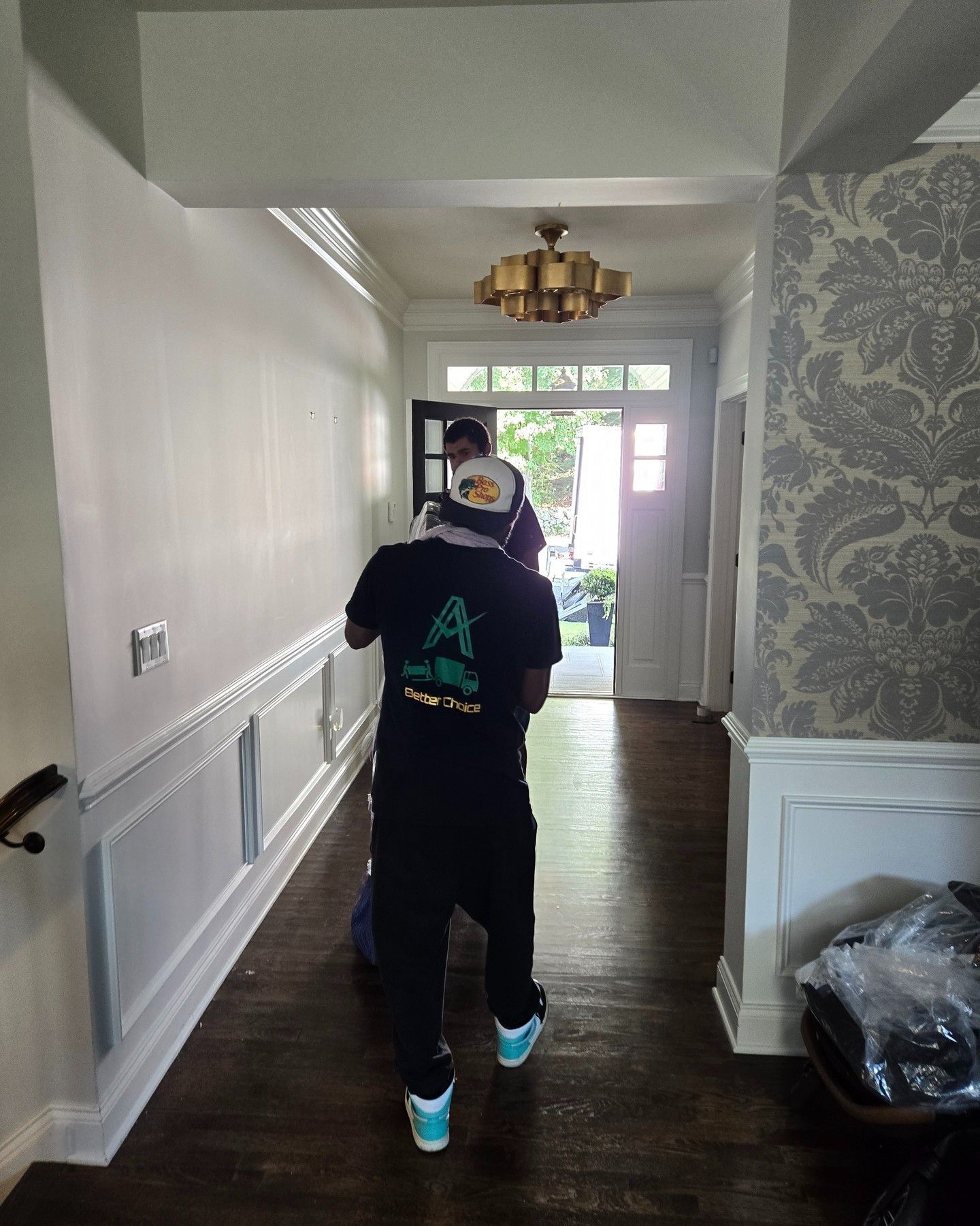 People in a hallway, entering a building. One person wears a black t-shirt and blue shoes. The walls are white and patterned.