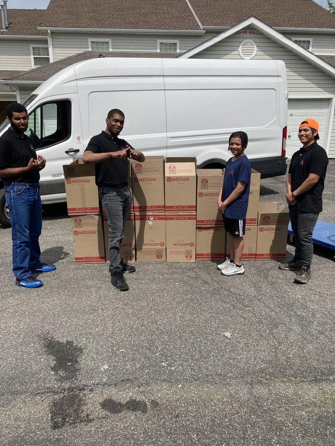 Four people standing with boxes in front of a white van on a driveway.