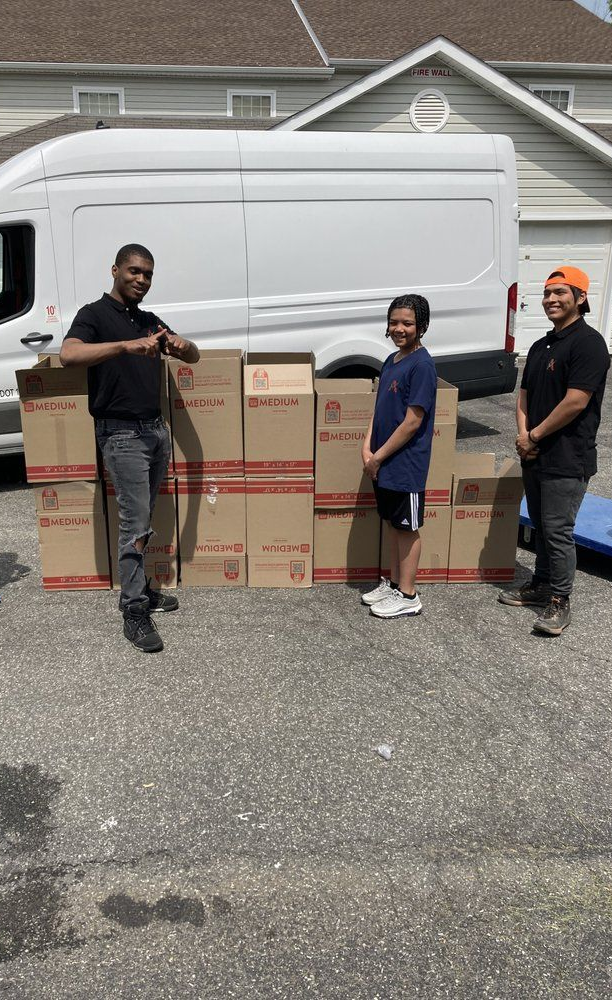 Four people standing with boxes in front of a white van on a driveway.