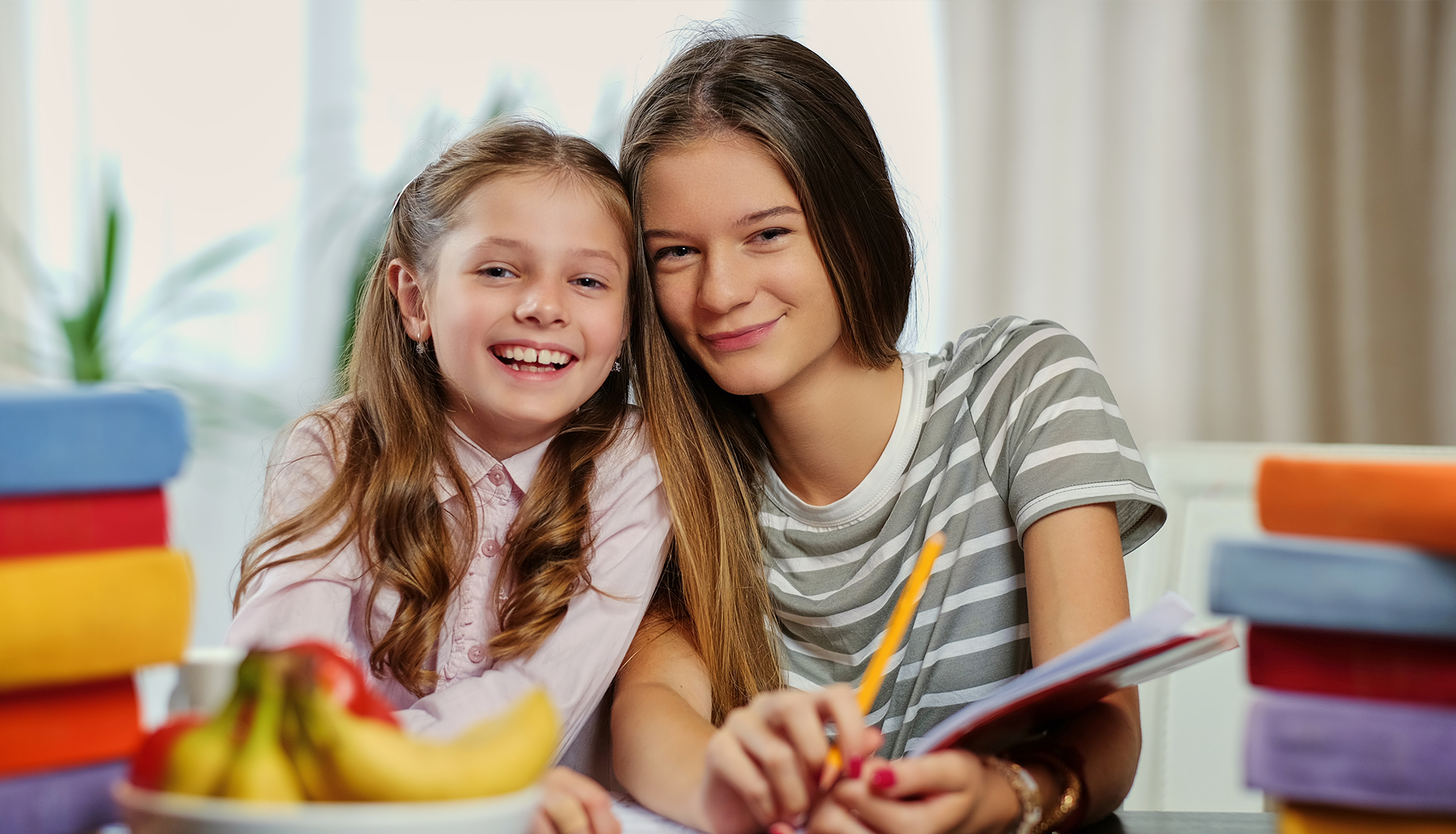 Two girls smiling at the camera, leaning close together at a table with books and fruit.