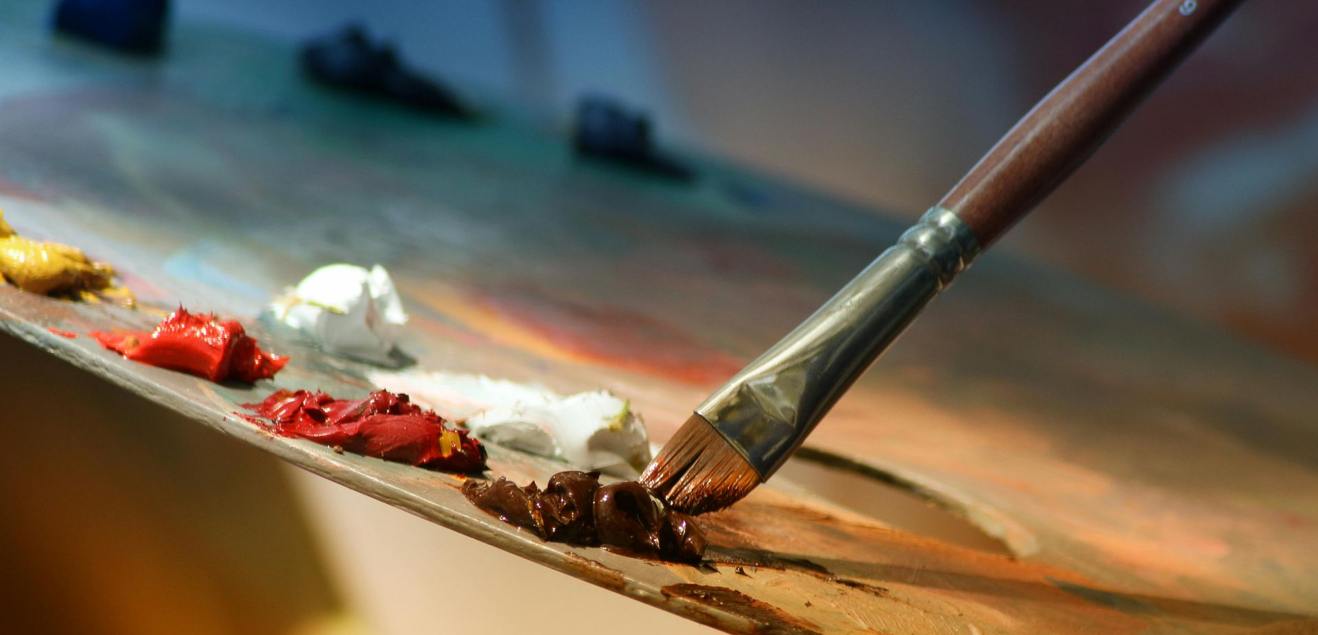Paintbrush mixing brown paint on a wooden artist's palette with various colored paint blobs.