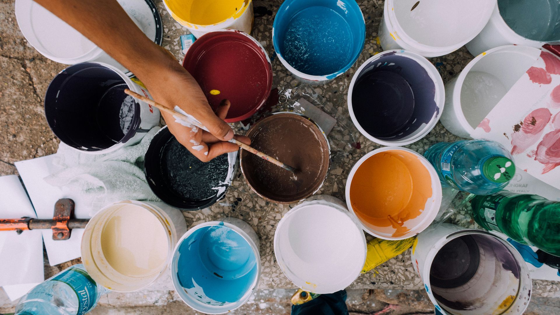 Person stirring brown paint in a bucket, surrounded by open paint buckets of various colors.