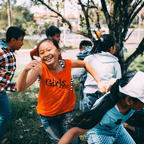 Children playing outdoors, one smiling in an orange shirt. Others are chasing each other near a tree.