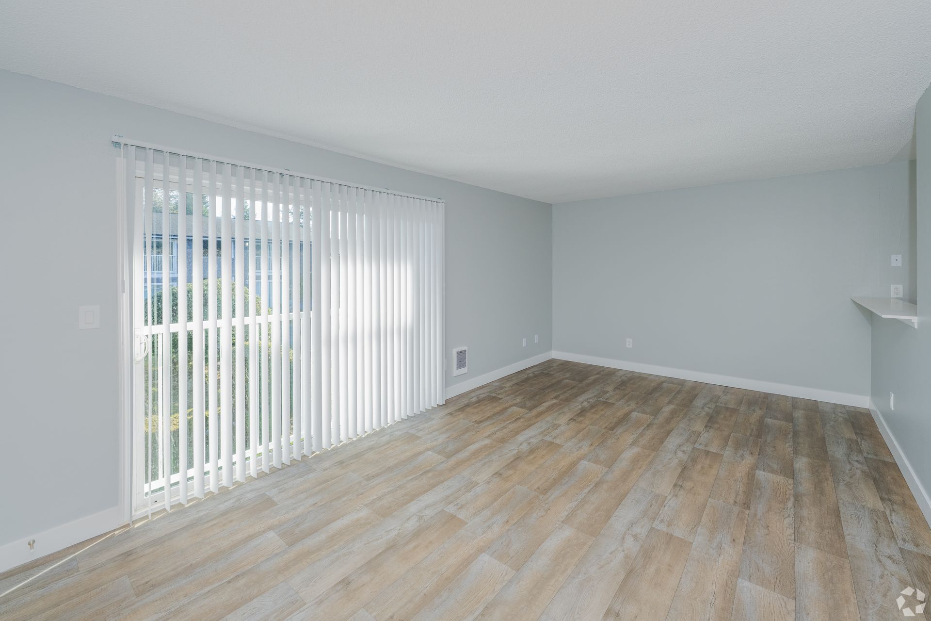 An empty room with light gray walls, wood-look flooring, and a large sliding glass door covered by white vertical blinds.
