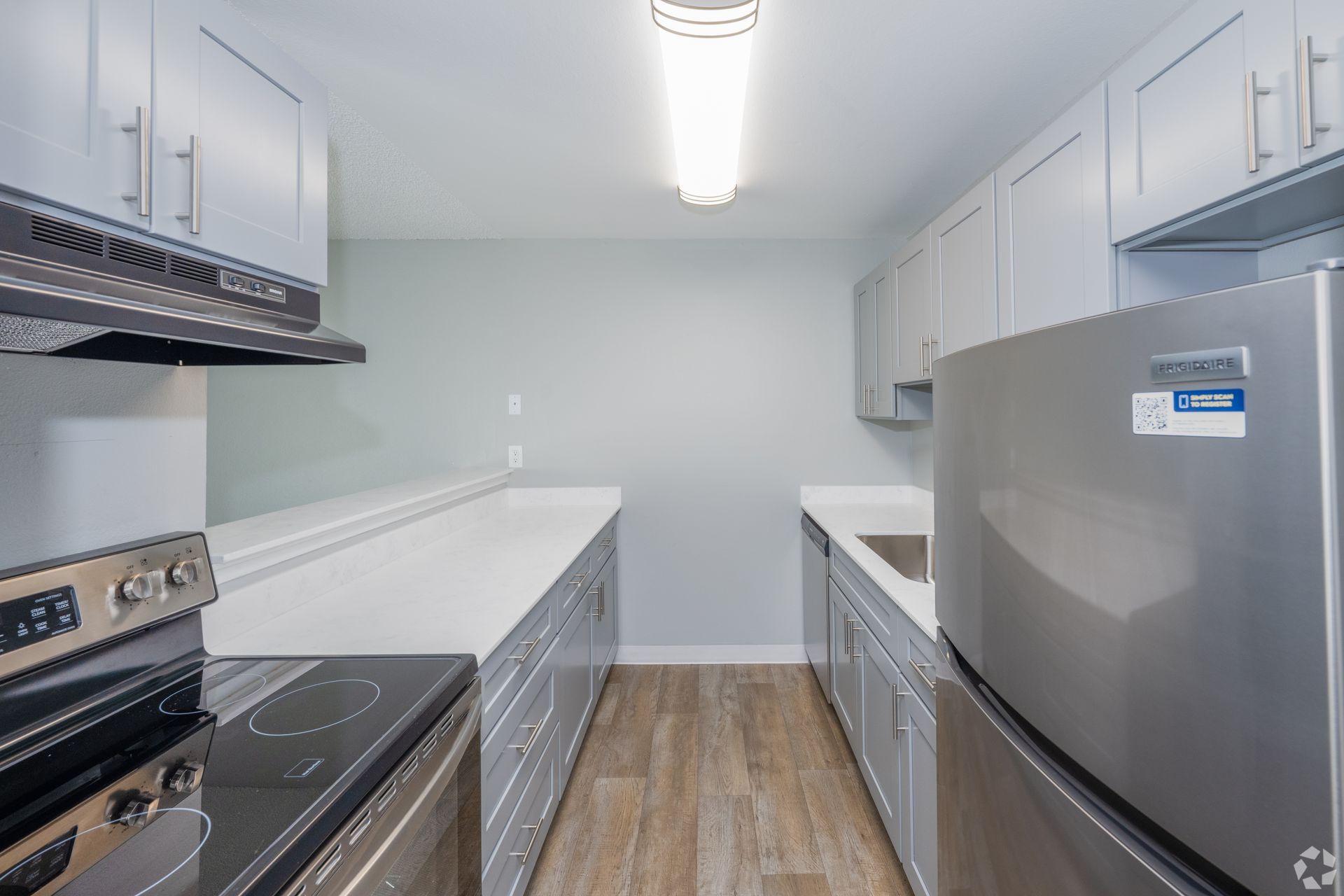 A galley-style kitchen with light gray cabinets, white countertops, stainless steel appliances, and wood-look flooring.