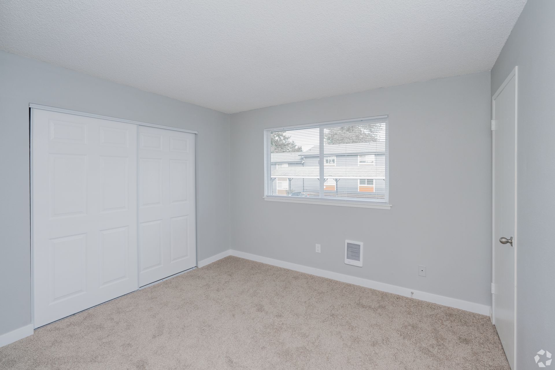 An empty bedroom with light gray walls, white sliding closet doors, beige carpeting, and a window overlooking a street.