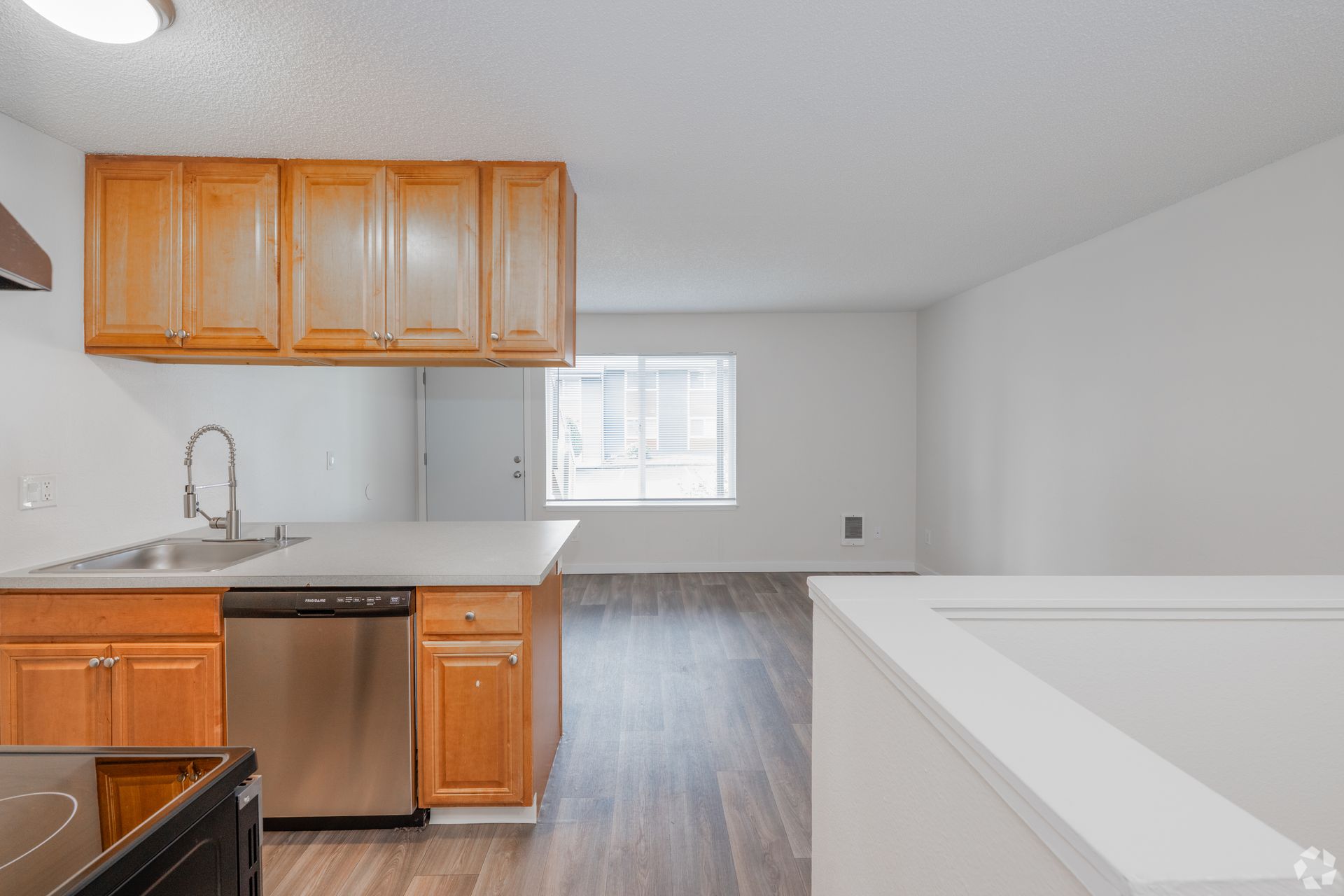 A view of a kitchen with wooden cabinets and stainless steel appliances opening into a sunlit, unfurnished living area.