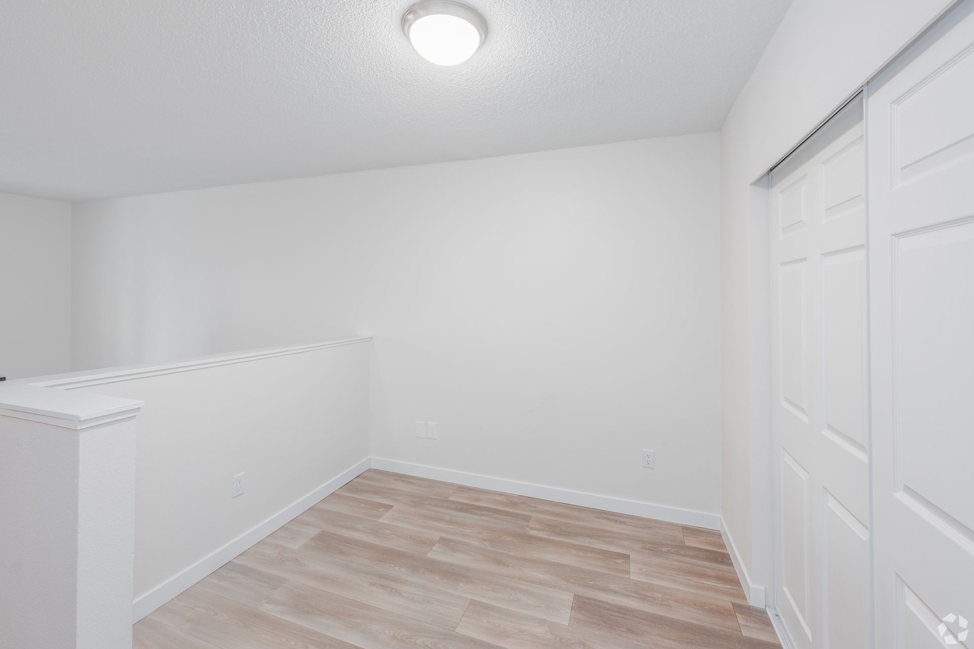 An empty, brightly lit residential landing area featuring light wood flooring, white walls, and a white closet door.