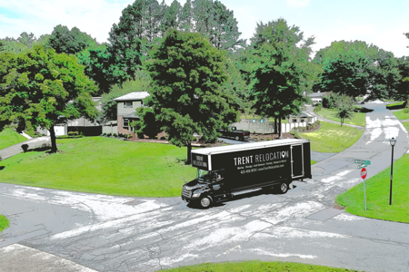 Truck turning at a tree-lined road intersection. The truck is black with white lettering. Green grass and houses in the background.