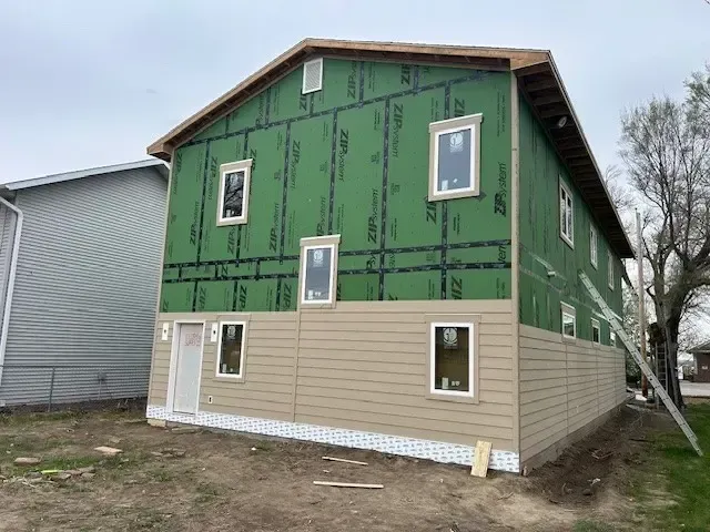 Two-story building under construction; green sheathing above tan siding, windows installed, unfinished roof.