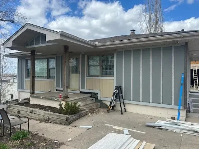 House exterior with gray and tan siding under construction, blue level, ladder, overcast sky.