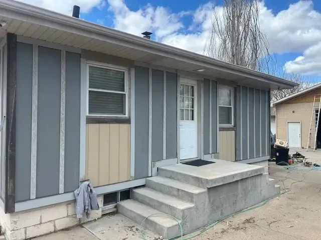 Exterior of a house with gray and tan siding, a concrete porch, and a white door under a cloudy sky.