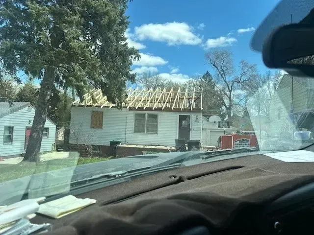 A house under construction with exposed roof rafters, viewed from inside a car on a sunny day.